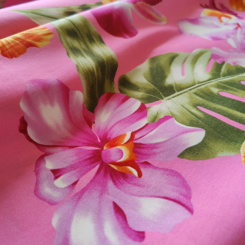 Tissu tahitien en coton, gros plan sur motifs orchidées et hibiscus orange et violet sur fond rose. Aucun réassort. Vendu en coupon.