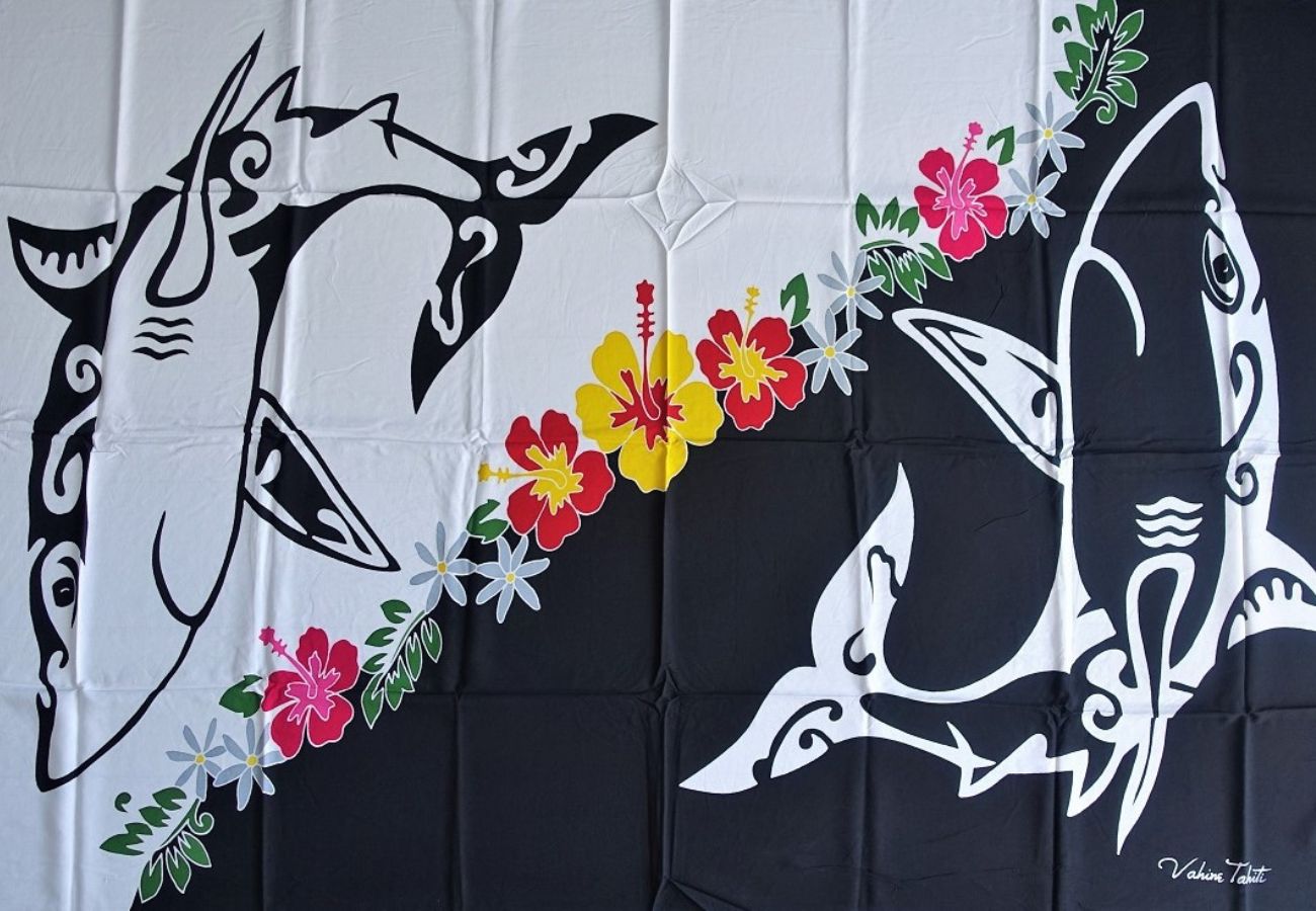 Paréo en fibranne représentnat des requins polynésiens noir et blanc séparés par une couronne de fleurs tropicales. 175 x 115 cm